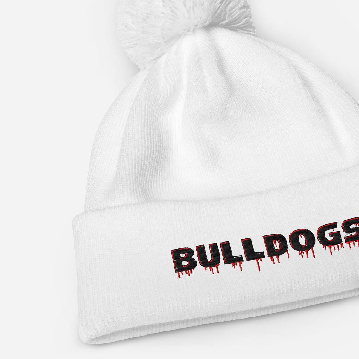 A white Bulldogs-inspired pom pom beanie featuring bold red-and-black embroidered lettering, soft-touch acrylic, a double-layer knit, and classic cuffed styling.