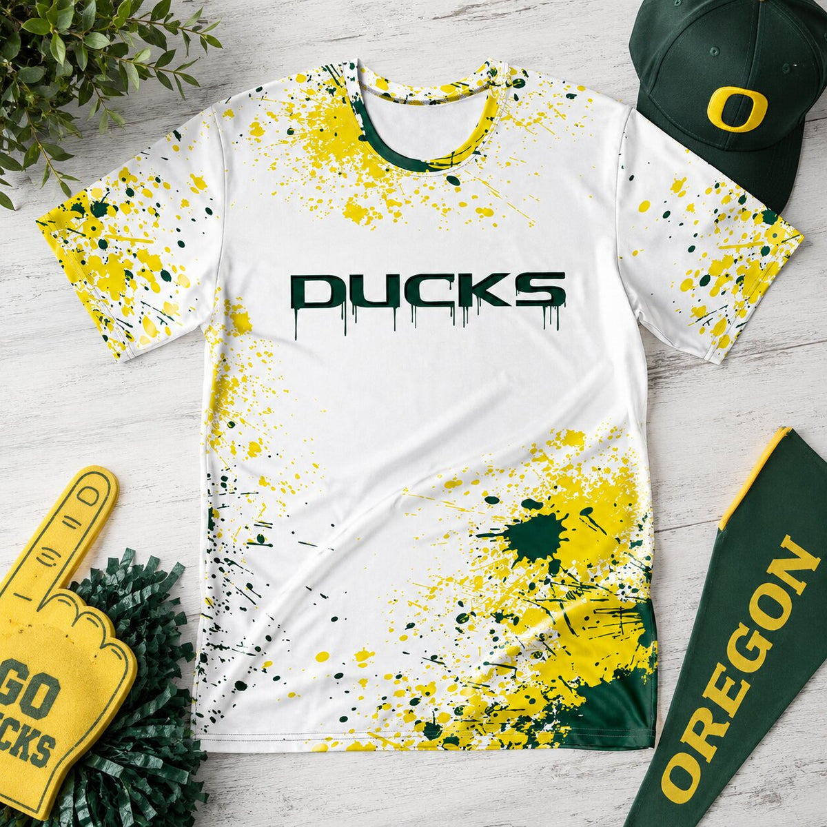 White Oregon Ducks paint splatter T-shirt laid flat on a light wood surface, styled with green-and-gold game day accessories including a Ducks cap, pom-poms, foam finger, and Oregon pennant.