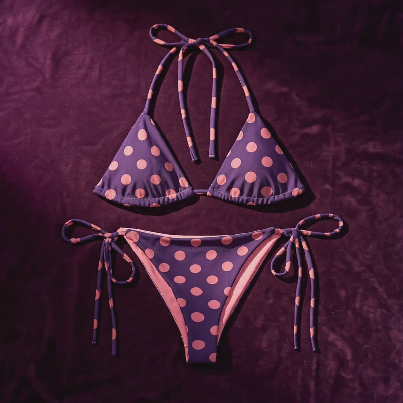 Female model wearing a purple and pink polka dot bikini poses on a cruise ship deck at golden hour with ocean views.